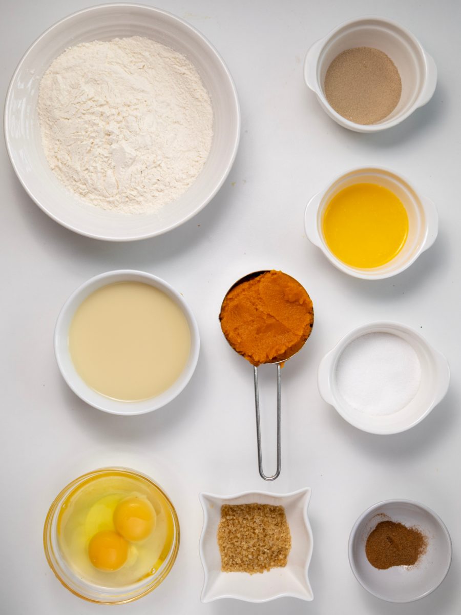 A flat lay shot of the ingredients for the pumpkin cinnamon rolls, including flour, milk, eggs, pumpkin puree, brown sugar, granulated sugar, and spices, arranged in white bowls on a white background.