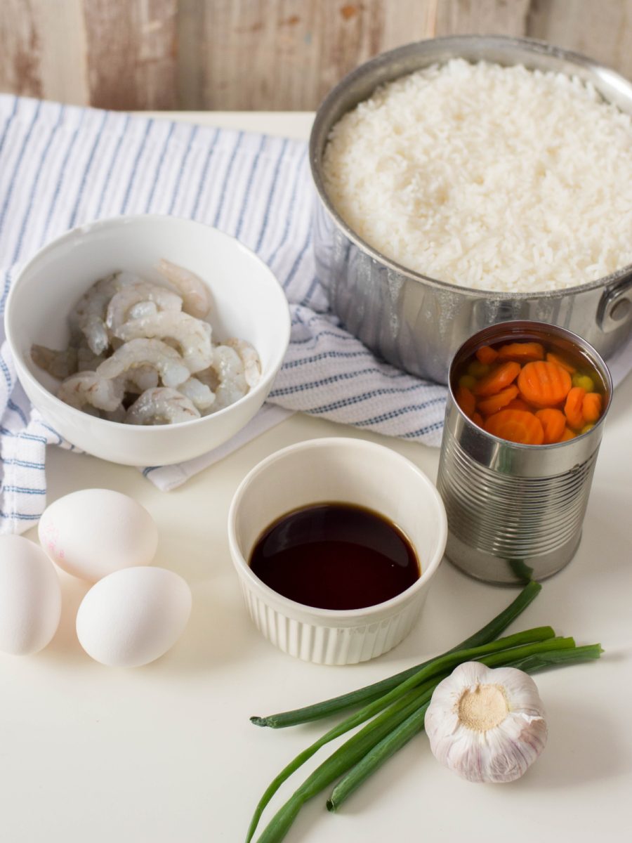 A vertical shot of the key ingredients for shrimp fried rice, including a pot of cooked rice, a can of peas and carrots, a bowl of raw shrimp, eggs, garlic, and soy sauce, all laid out on a white surface.