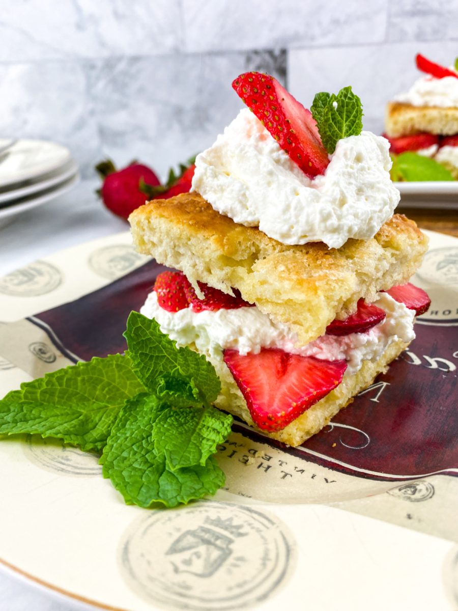 A beautifully styled, close-up shot of a strawberry shortcake on a decorative plate with a sprig of fresh mint. The shortcake is layered with fresh strawberries and whipped cream. In the background, out of focus, are more strawberries and a stack of plates.