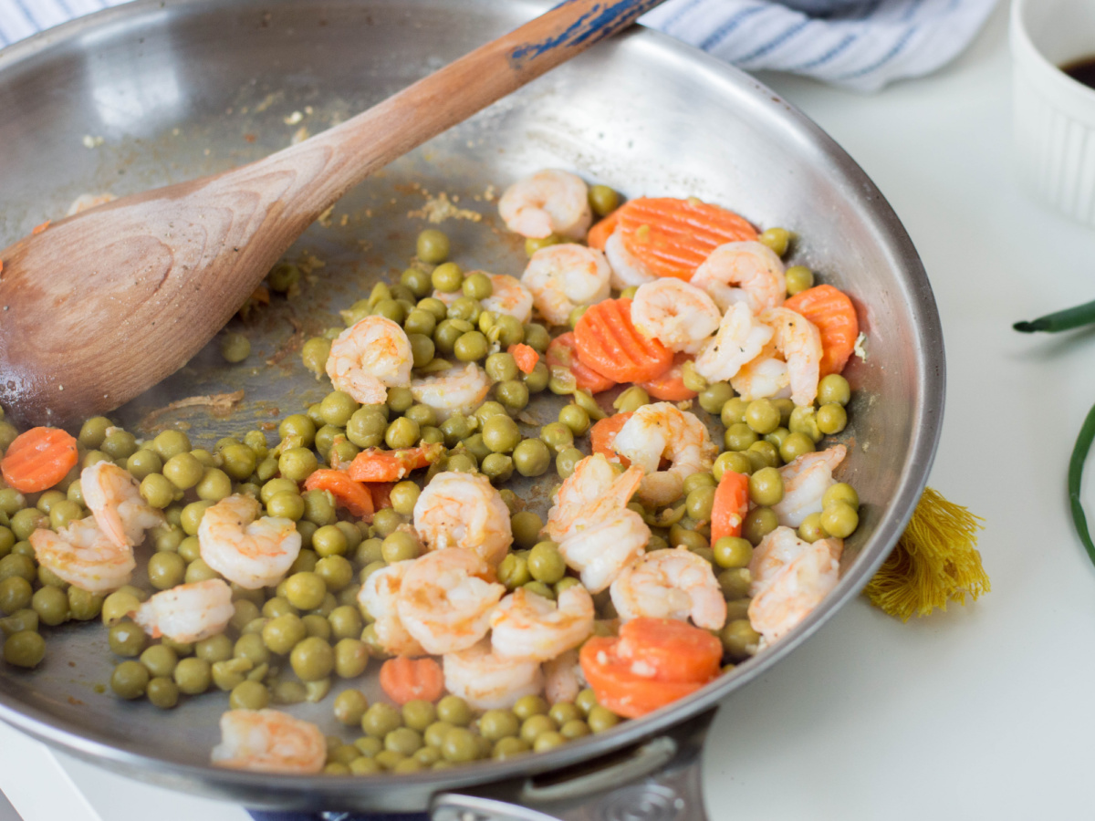 A stainless steel skillet filled with plump shrimp, bright green peas, and sliced carrots cooking together.