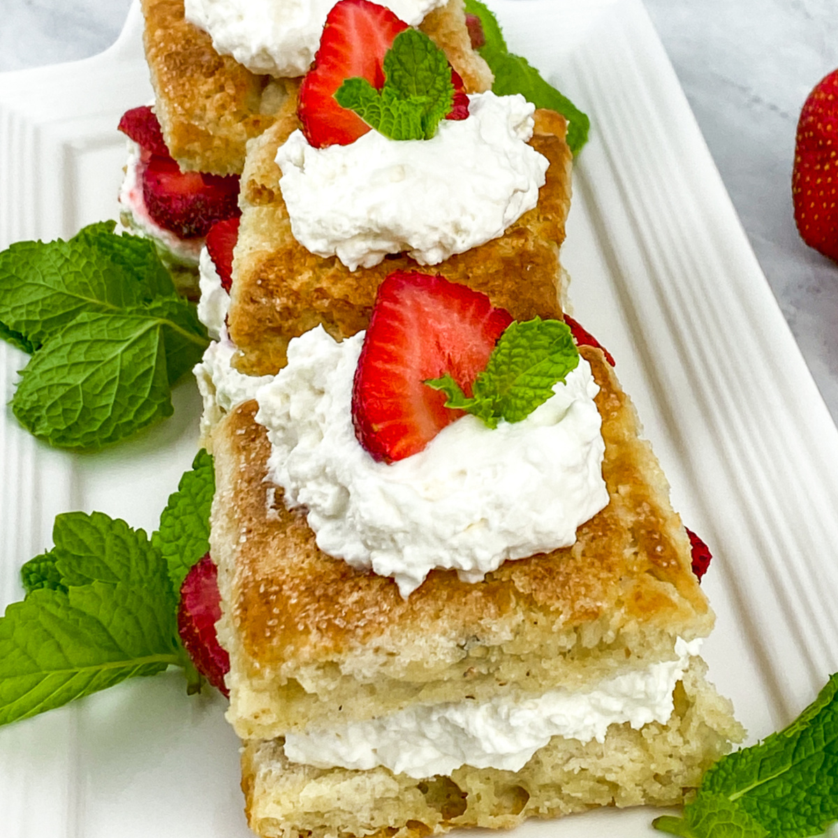 Three homemade strawberry shortcakes lined up on a long, rectangular white plate. Each shortcake is made with a sweet biscuit, layered with whipped cream and fresh strawberries, and garnished with a mint leaf. The shortcakes are viewed from a slightly elevated angle.