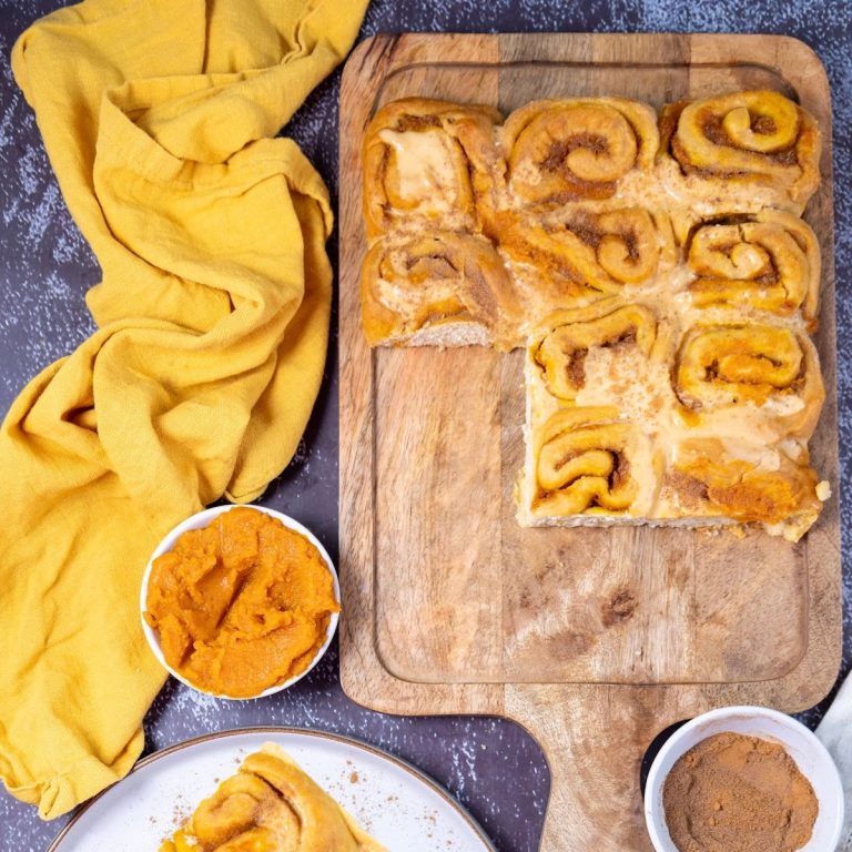 A feature image for a blog post showing pumpkin cinnamon rolls with cream cheese frosting on a white platter, with a pumpkin and kitchen tools in the background.