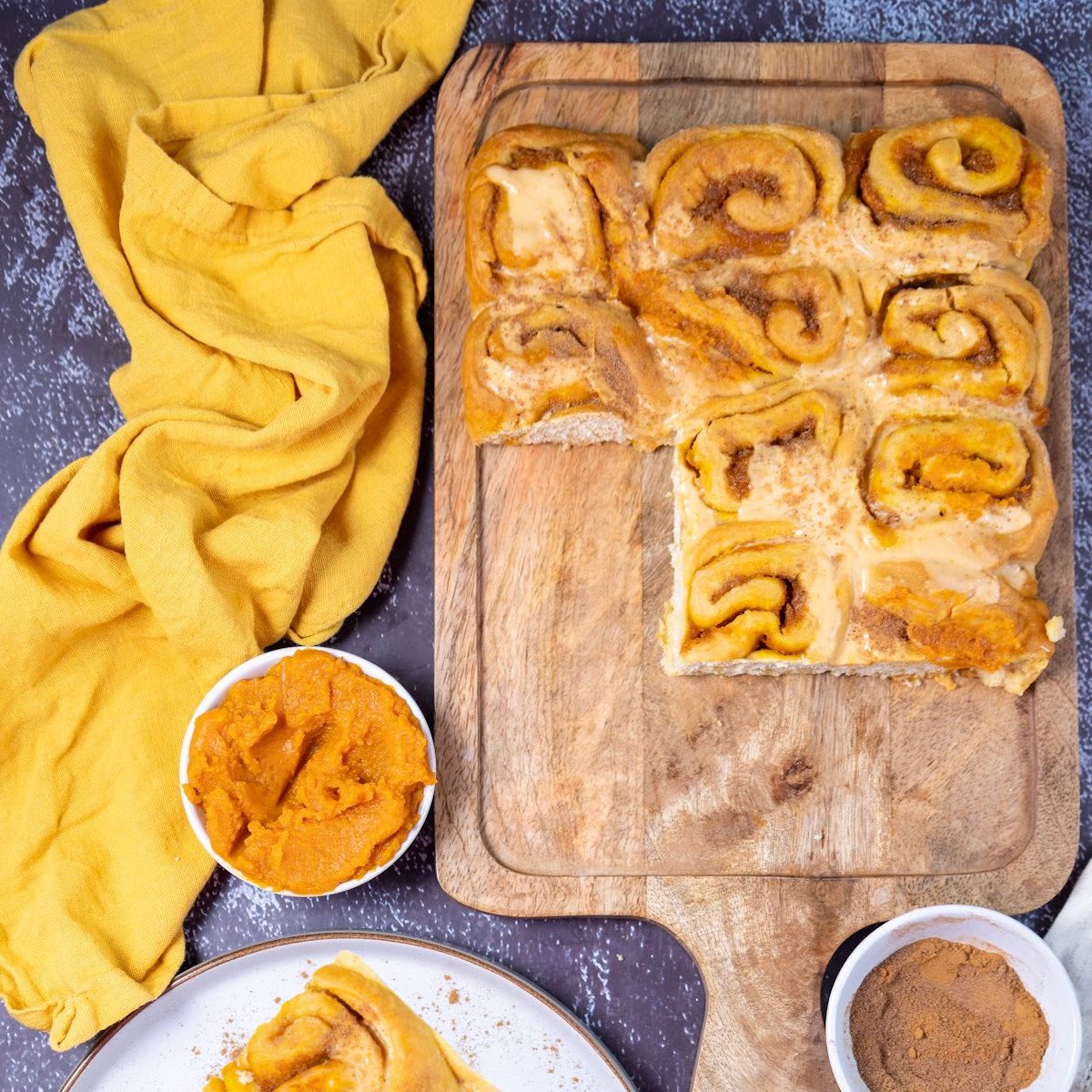 A feature image for a blog post showing pumpkin cinnamon rolls with cream cheese frosting on a white platter, with a pumpkin and kitchen tools in the background.