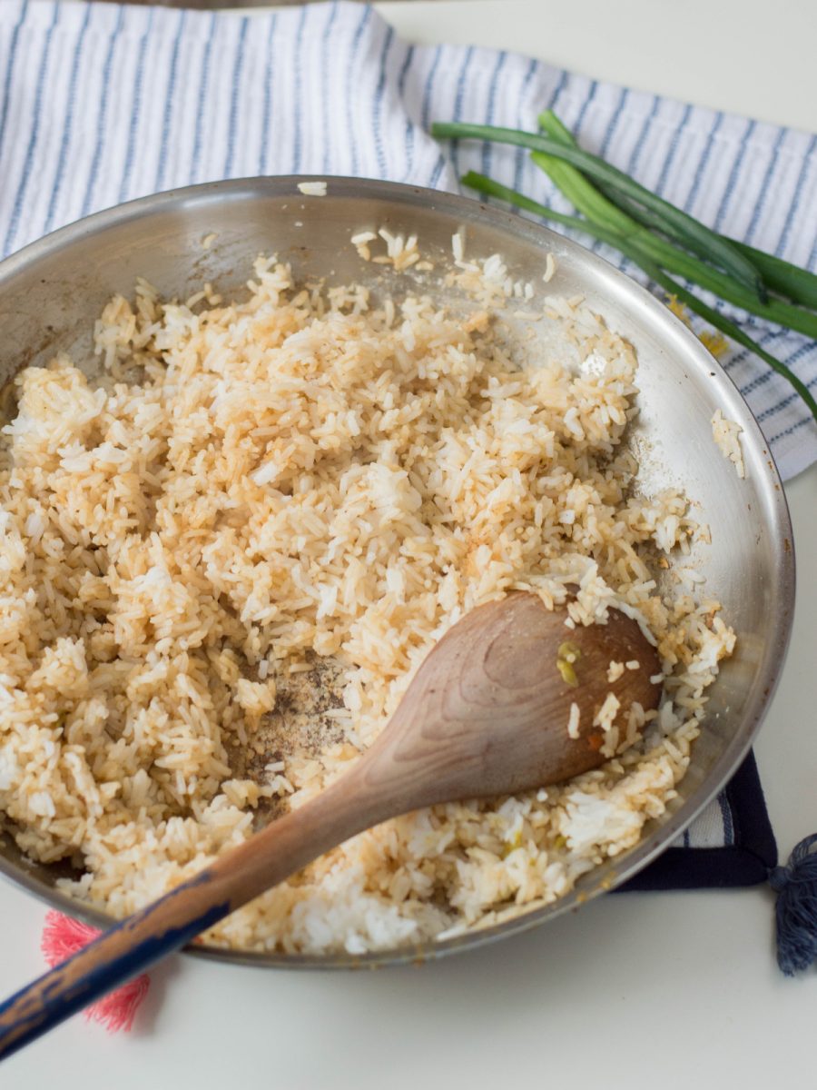 Cooked white rice is being stirred in a skillet with a wooden spoon, coated in soy sauce and other seasonings.