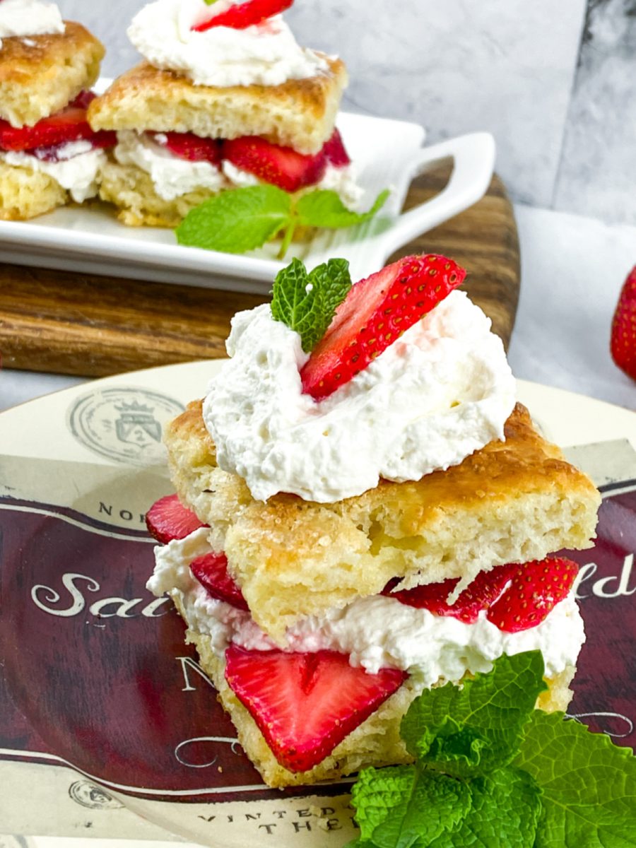 A close-up, eye-level shot of a homemade strawberry shortcake on a decorative plate, garnished with a sprig of fresh mint. The shortcake is made with two biscuit halves, layered with whipped cream and sliced strawberries. Another shortcake is visible in the background.