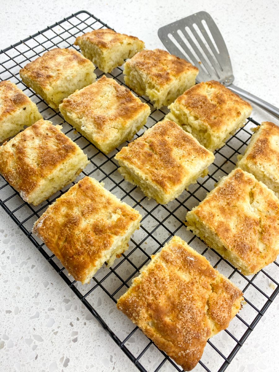 Freshly baked, square-shaped biscuits on a wire cooling rack. The biscuits have a golden-brown top dusted with a light layer of raw sugar. A metal spatula is resting on the right side of the rack.