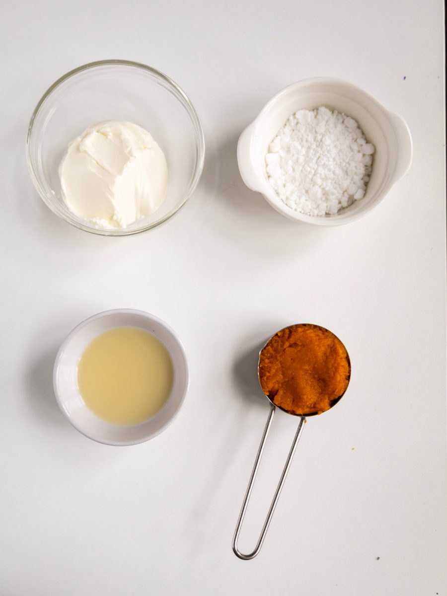 A flat lay shot of the four ingredients for the frosting, including cream cheese, powdered sugar, pumpkin puree, and milk, arranged in small white bowls on a white background.