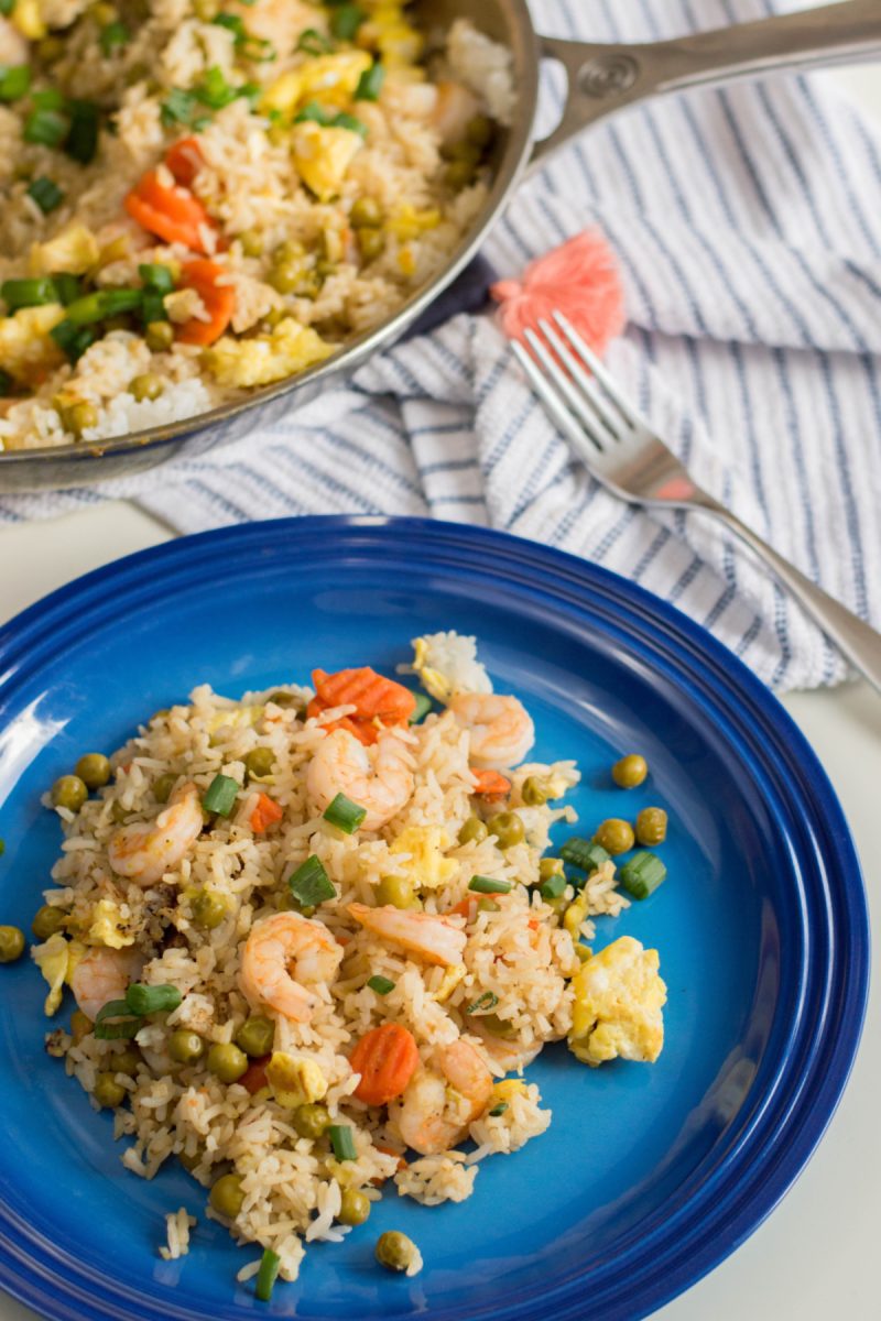 An overhead view of a blue plate with a serving of shrimp fried rice, with a skillet of the finished dish and a fork in the background.