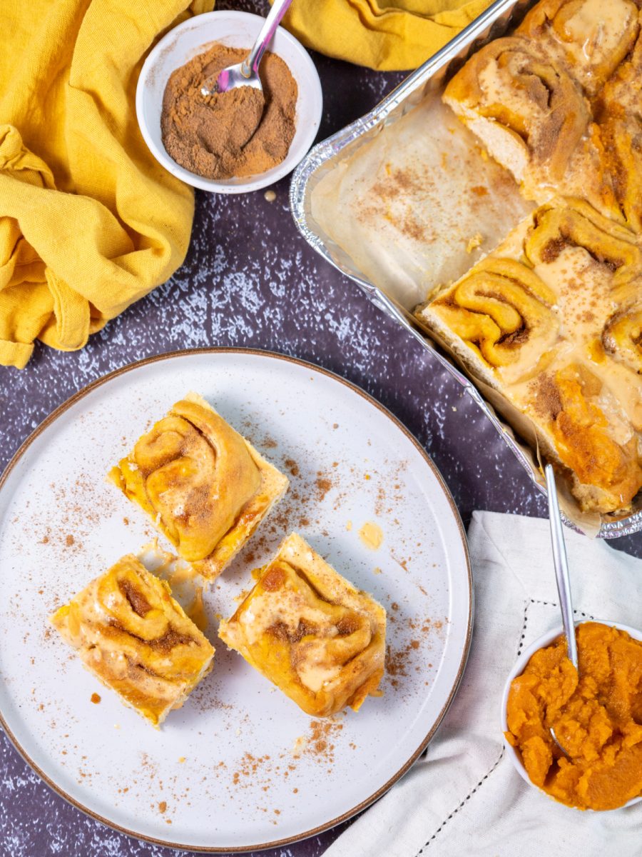 An overhead view of a baking pan of baked pumpkin cinnamon rolls next to a bowl of pumpkin puree, with a sprinkling of spices.