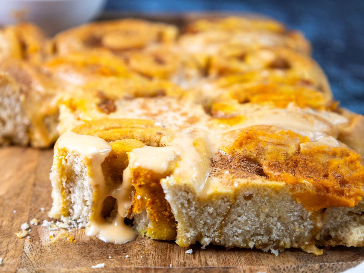 A close-up shot of several frosted pumpkin cinnamon rolls arranged on a rustic wooden cutting board, with a sprinkle of cinnamon on top.