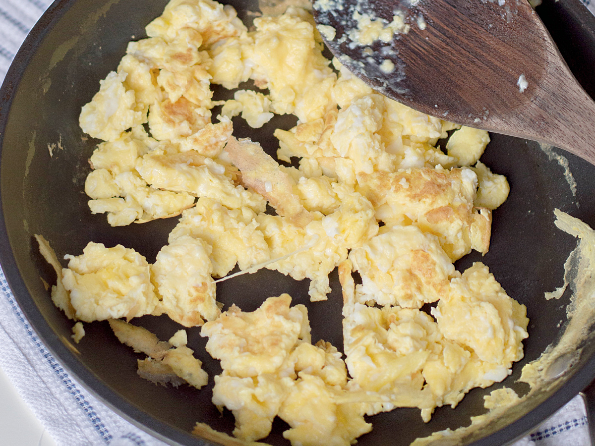 A close-up shot of fluffy scrambled eggs being cooked in a non-stick skillet with a wooden spatula.