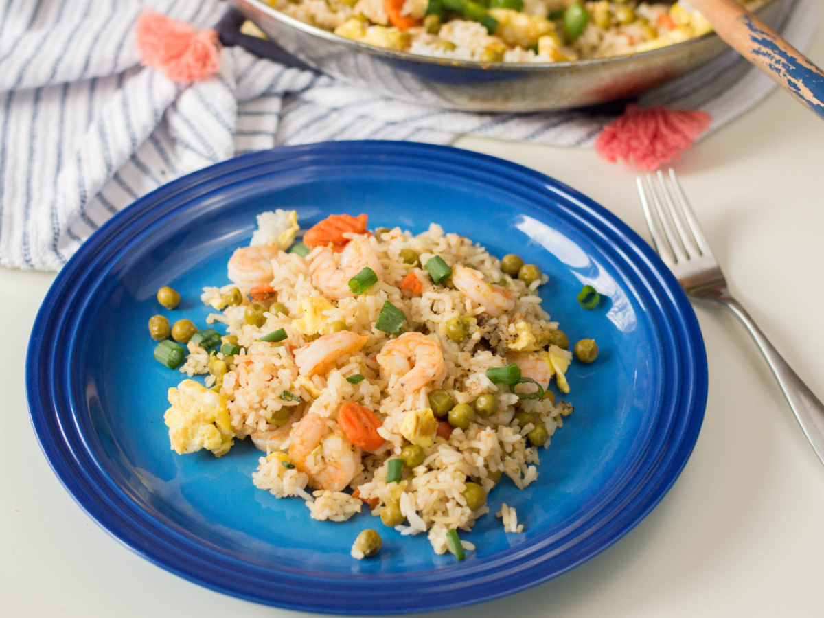 A blue plate holding a serving of shrimp fried rice sits on a white table, with a skillet containing more fried rice visible in the background.