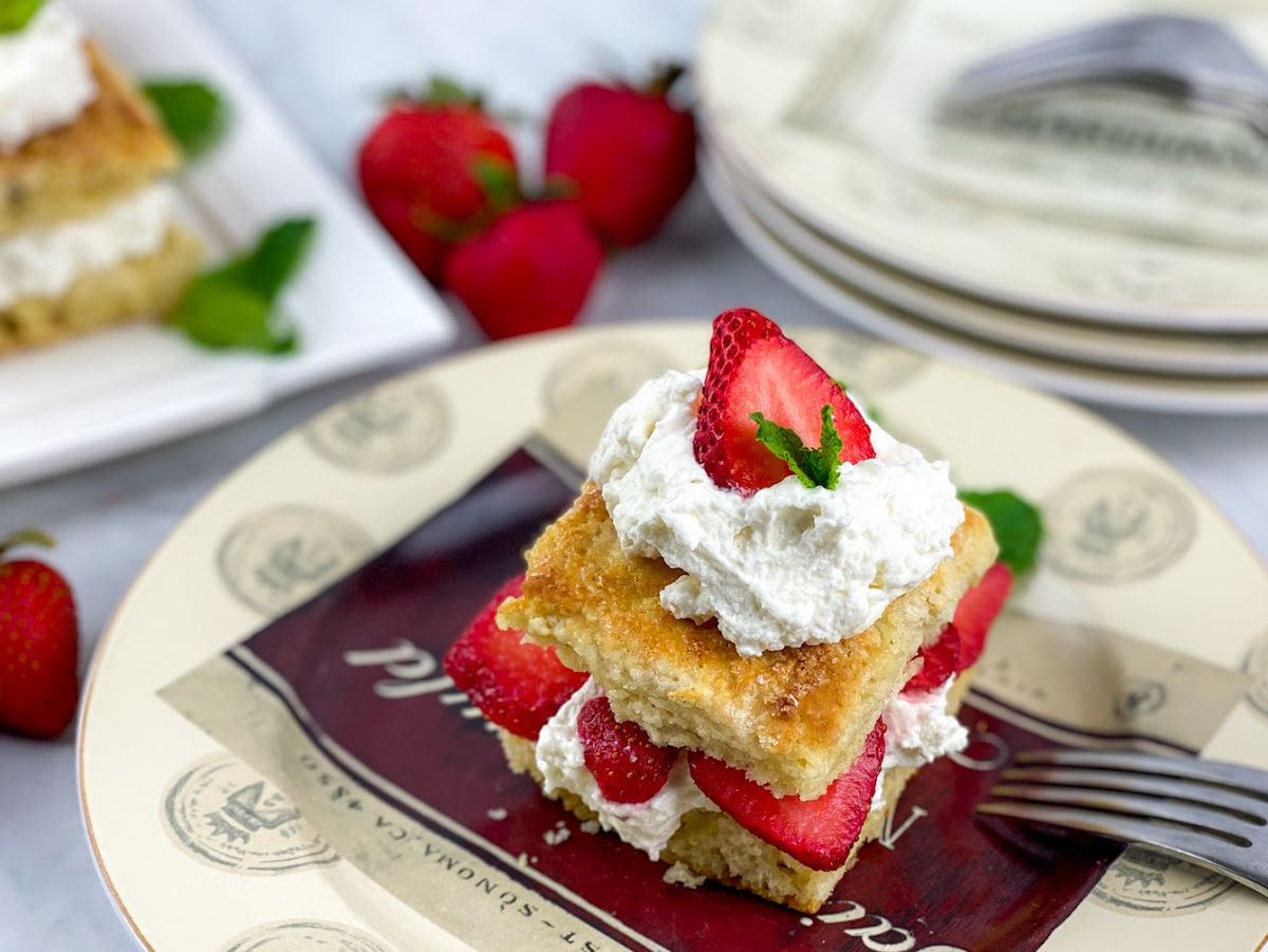 A close-up shot of a homemade strawberry shortcake on a decorative plate. The shortcake is made of two biscuit halves, filled with whipped cream and sliced strawberries, and topped with more cream, a strawberry slice, and a mint leaf. A fork is visible on the plate, and more shortcakes and plates are in the soft-focus background.