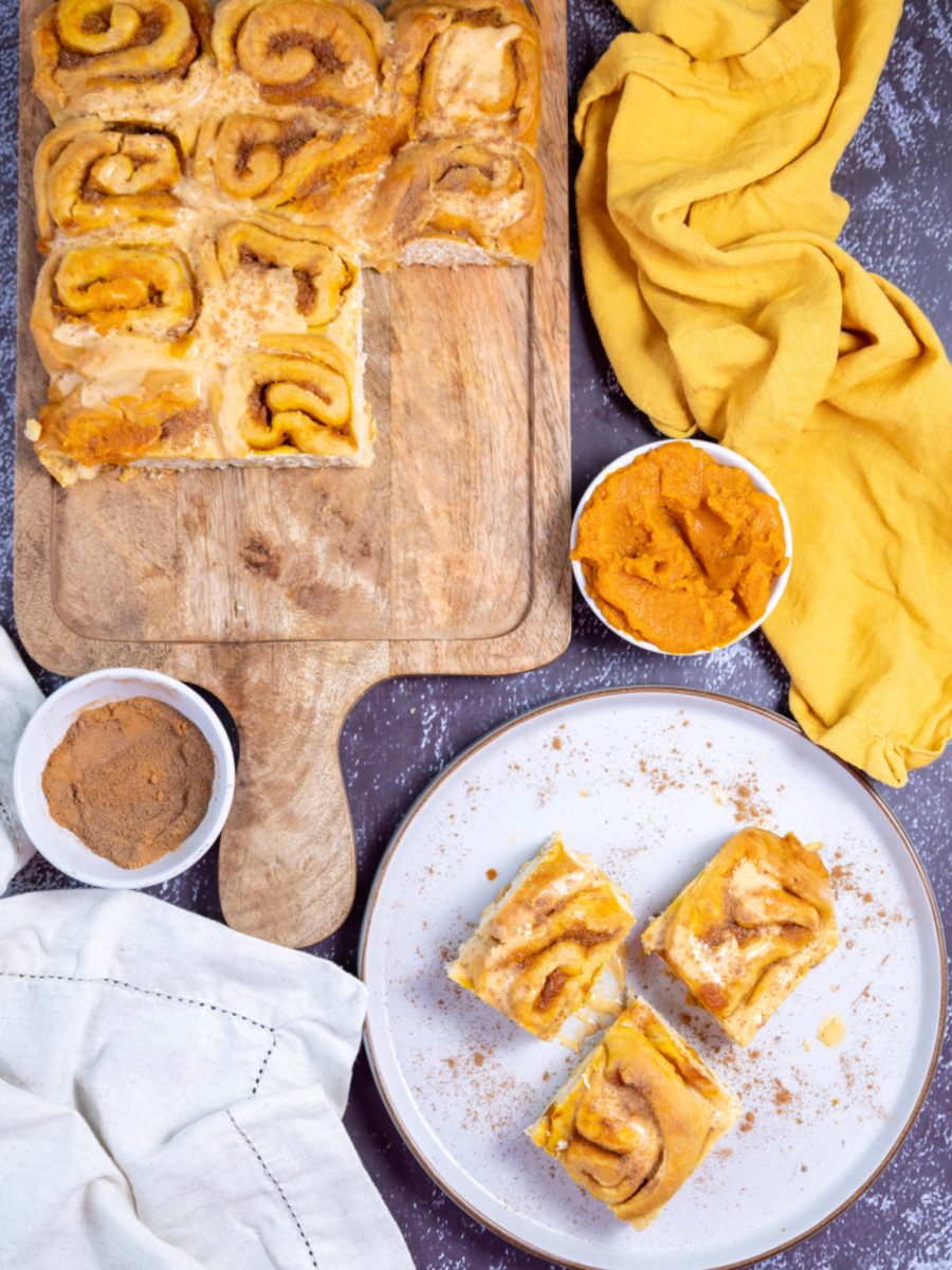 A scene showing a wooden board with freshly baked cinnamon rolls and a plate holding three rolls, all topped with a creamy frosting.