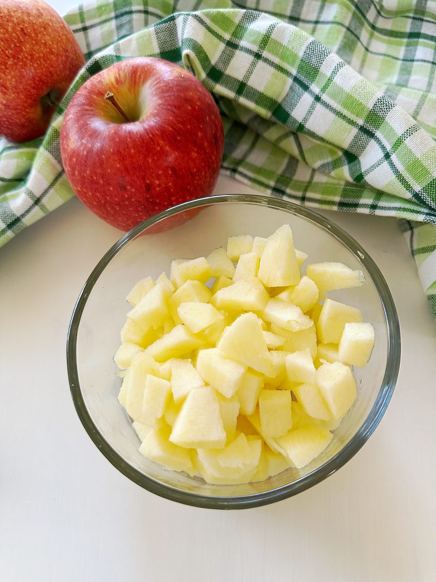 A clear glass bowl filled with freshly diced apples on a white surface, with a green plaid towel and two whole red apples in the background.