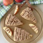 A close-up shot of three glazed apple chai scones on a wooden cutting board, with apple slices and cinnamon sticks scattered around them.