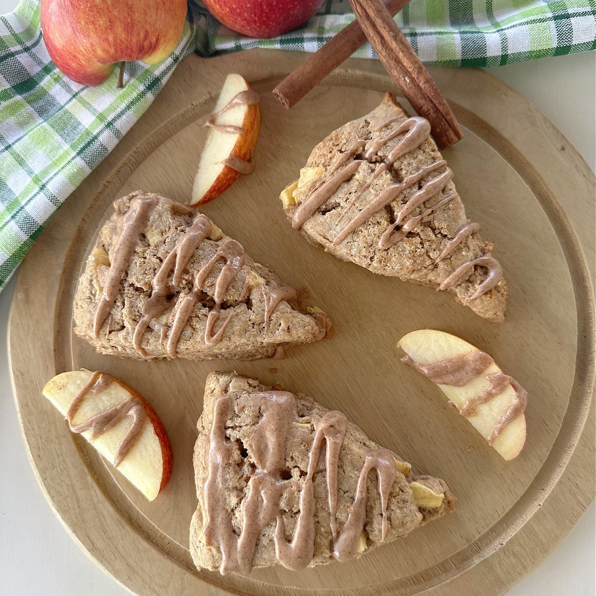 A close-up shot of three glazed apple chai scones on a wooden cutting board, with apple slices and cinnamon sticks scattered around them.