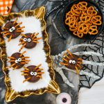 Overhead view of five finished Halloween Spider Donuts on a gold and white serving tray, with a bowl of pretzels, a skeleton hand, and orange and black fabric visible on a marble surface.