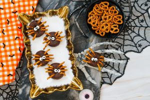 Overhead view of five finished Halloween Spider Donuts on a gold and white serving tray, with a bowl of pretzels, a skeleton hand, and orange and black fabric visible on a marble surface.