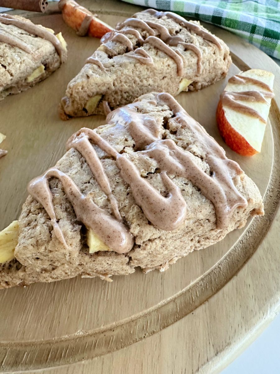 A close-up, low-angle shot of a single baked apple chai scone drizzled with glaze, with two other scones and apple slices blurred in the background on a wooden board.