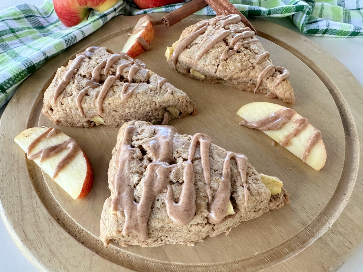 An overhead shot of three apple chai scones with a glaze drizzle on a round wooden board. The scones are surrounded by apple slices, cinnamon sticks, and two fresh apples.