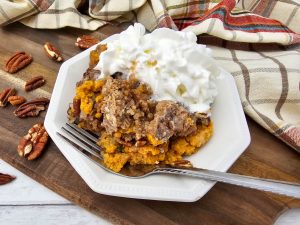 A white plate holding a serving of warm Crock Pot Pumpkin Dump Cake with crumble topping and pecans, covered in a mound of whipped cream, set on a wooden board next to a fall-themed plaid napkin and whole pecans.