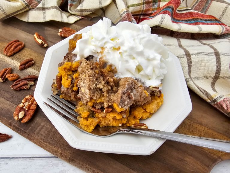 A white plate holding a serving of warm Crock Pot Pumpkin Dump Cake with crumble topping and pecans, covered in a mound of whipped cream, set on a wooden board next to a fall-themed plaid napkin and whole pecans.