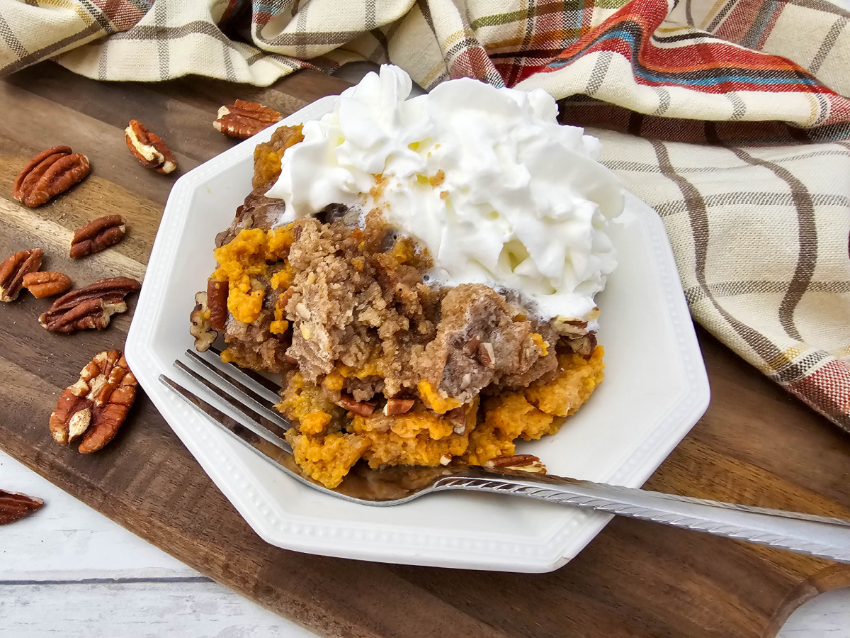 A white plate holding a serving of warm Crock Pot Pumpkin Dump Cake with crumble topping and pecans, covered in a mound of whipped cream, set on a wooden board next to a fall-themed plaid napkin and whole pecans.