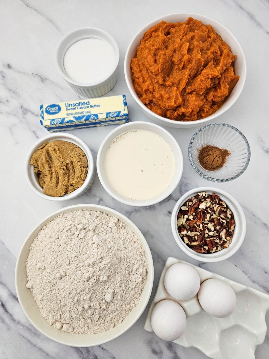 An overhead flat lay photo showing all measured ingredients for the Crock Pot Pumpkin Dump Cake, including a bowl of pumpkin puree, a bowl of spice cake mix, butter, eggs, evaporated milk, brown sugar, granulated sugar, pumpkin pie spice, and chopped pecans, all arranged on a white marble surface.