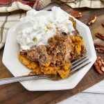 A close-up serving of warm Crock Pot Pumpkin Dump Cake on a white plate, featuring a creamy orange pumpkin custard base topped with a buttery, golden-brown crumble and pecans, finished with a generous dollop of whipped cream.