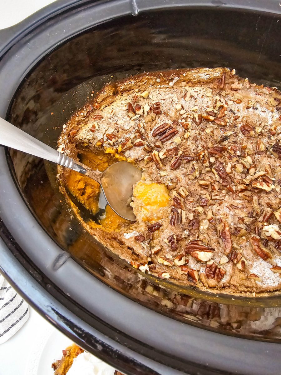 A close-up, overhead view of the finished Crock Pot Pumpkin Dump Cake inside the slow cooker insert, showing the golden-brown, buttery cake crumble topping and chopped pecans. A spoon scoops out a warm serving, revealing the creamy orange pumpkin custard underneath.