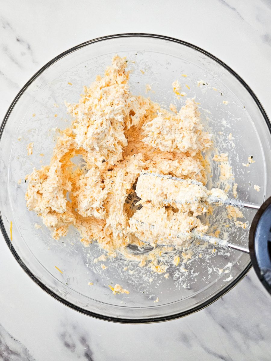 Close-up overhead view of a clear glass bowl containing a creamy, light orange cheese mixture being blended with an electric hand mixer. Specks of green seasoning are visible in the mixture.