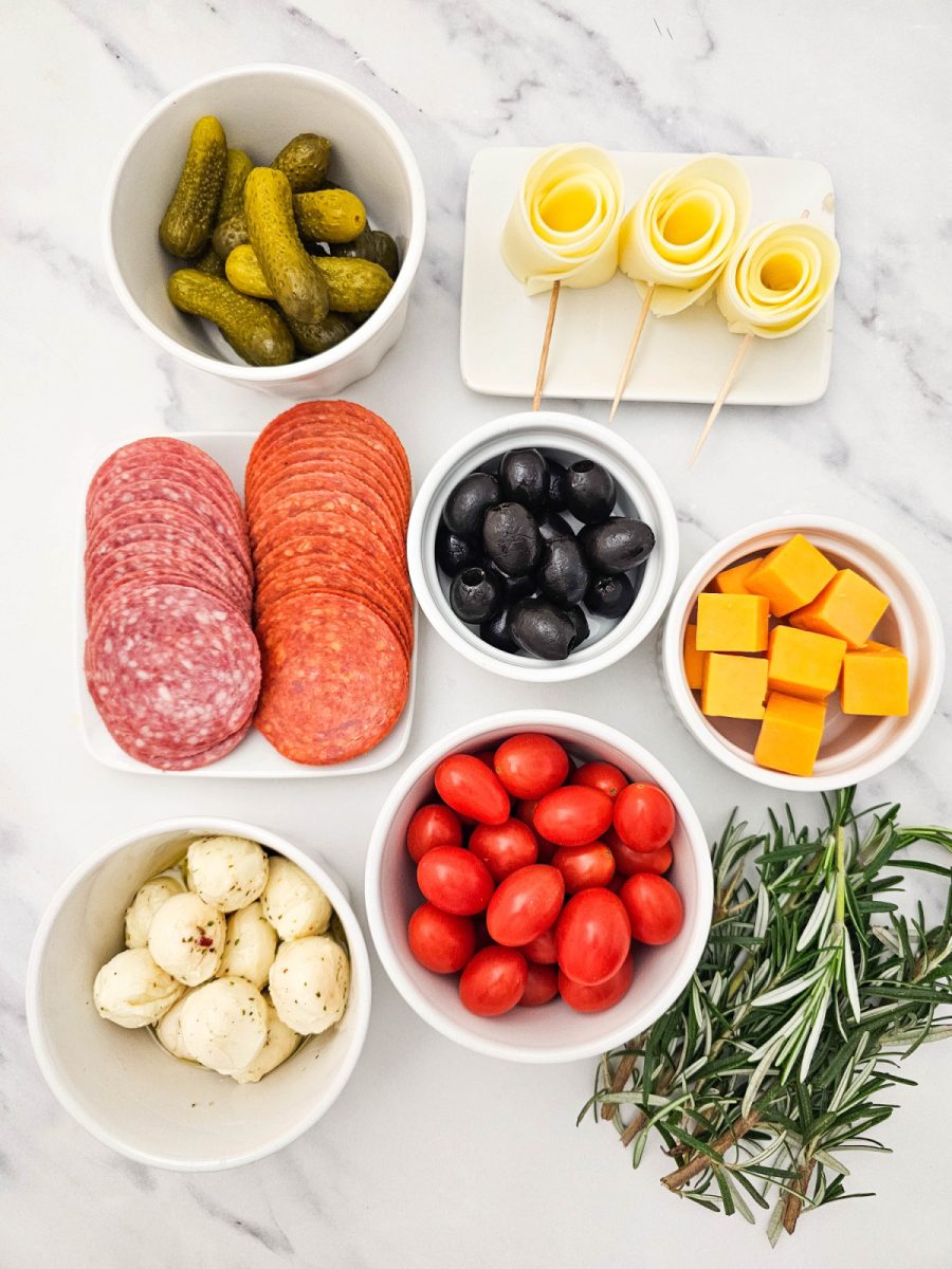 Overhead flat lay showing all the prepared ingredients for the charcuterie wreath: bowls of gherkins, black olives, cherry tomatoes, cubed cheddar, marinated mozzarella balls, stacks of salami and pepperoni, three provolone cheese roses, and a bunch of fresh rosemary.