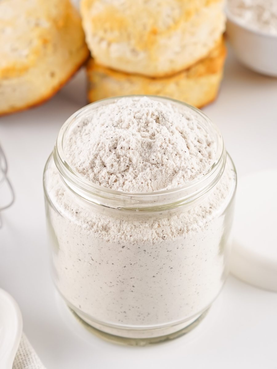 Close-up shot of a clear jar filled with the homemade, shelf-stable dry white gravy mix, featuring black pepper flakes visible in the flour and dry milk powder.