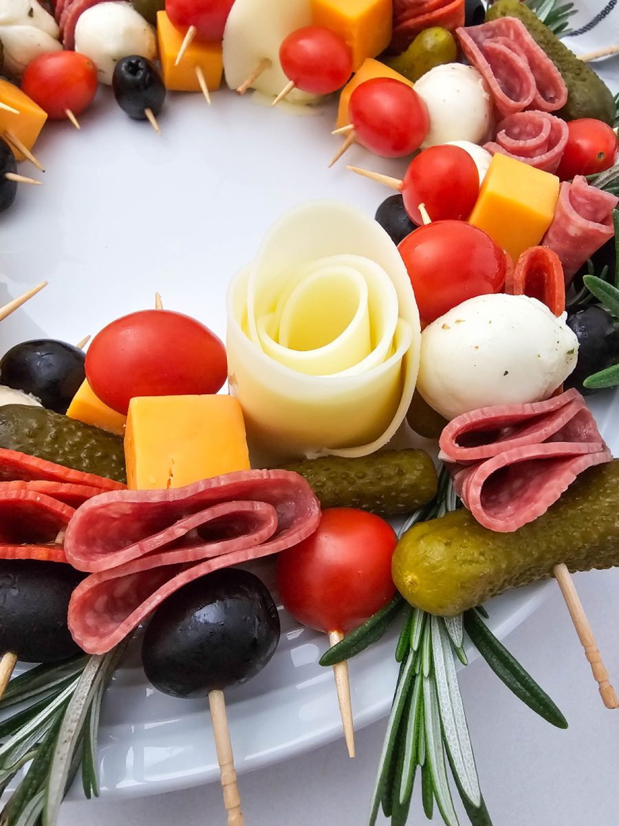 Macro close-up shot focusing on a Provolone cheese rose centerpiece, surrounded by olives, tomatoes, salami, cheddar cubes, and gherkins on the charcuterie wreath.