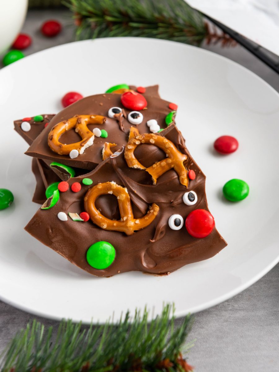 A close-up stack of broken Reindeer Chocolate Bark pieces on a white plate, showing the pretzel antlers, red M&M noses, candy eyes, and green M&M decorations.