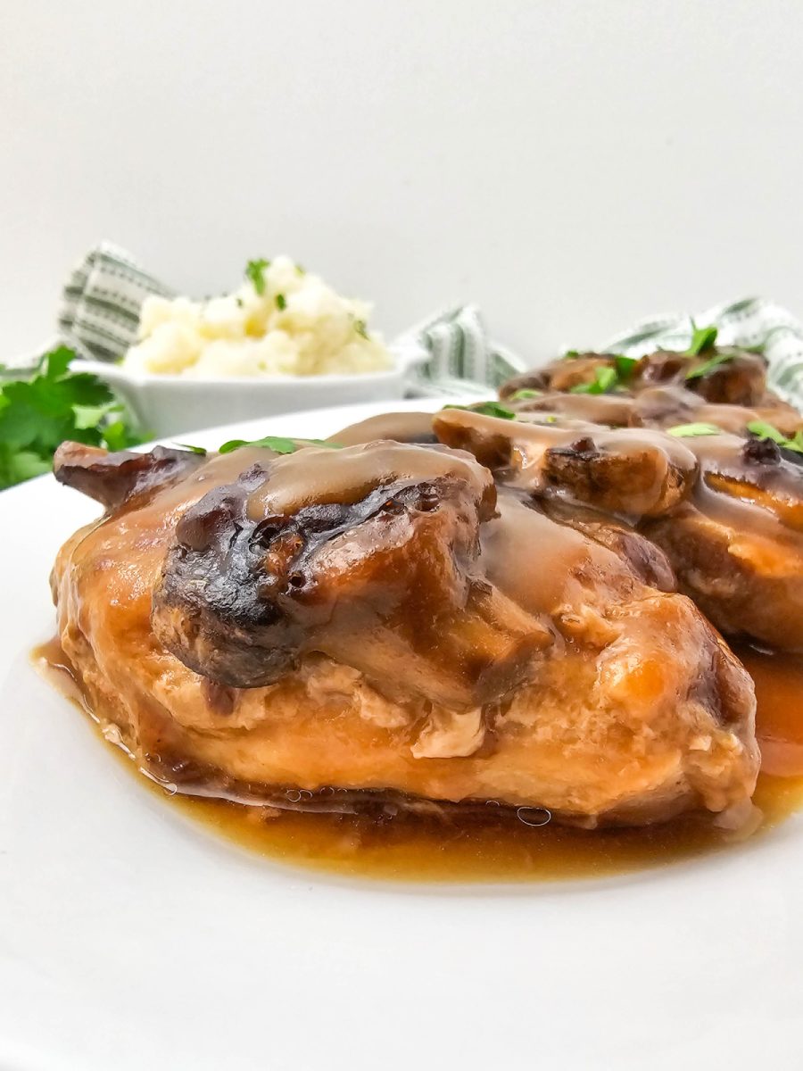 Extreme close-up of a single smothered pork chop showing the thick brown gravy and mushroom slices, with a blurred bowl of mashed potatoes in the background.