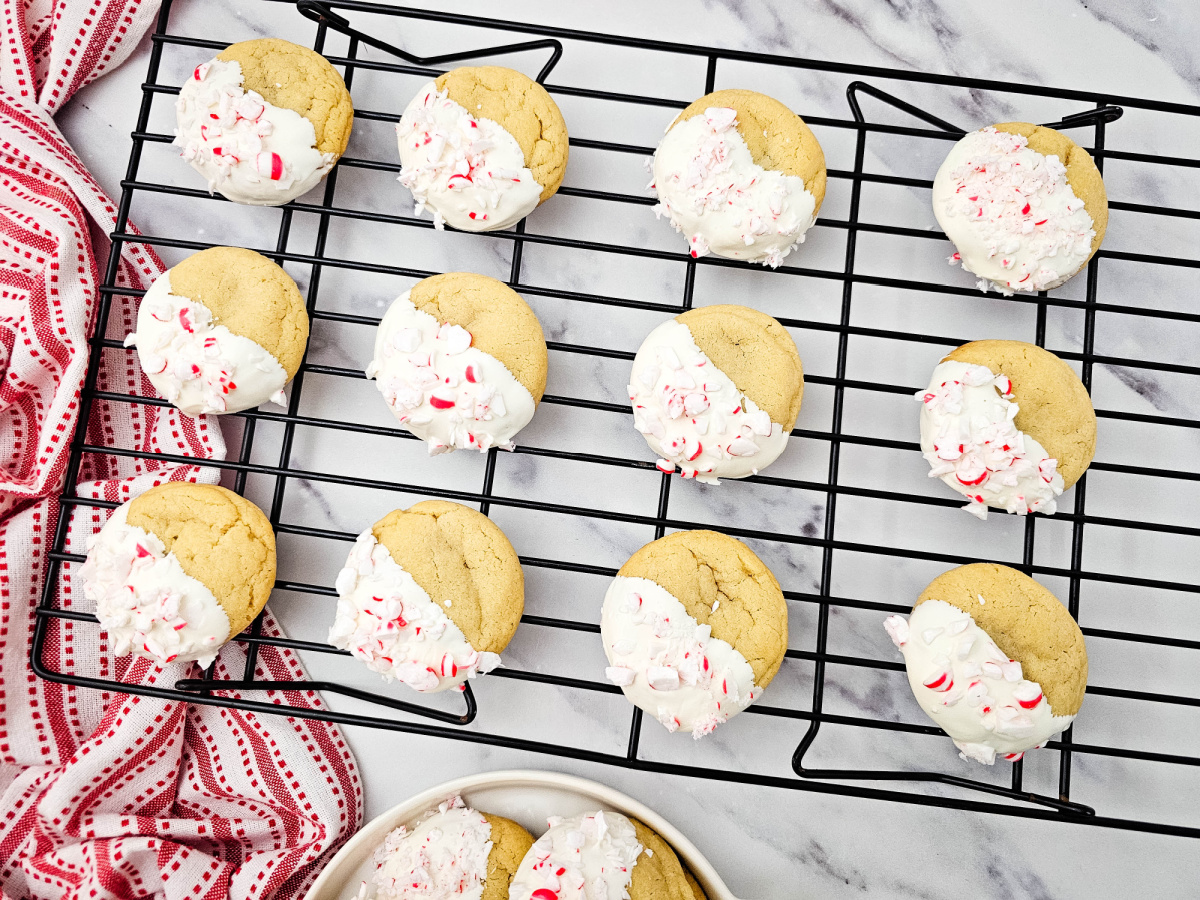 A dozen freshly baked Peppermint Crunch Cookies cooling on a black wire rack, showcasing their golden base and the vibrant white chocolate and peppermint crunch topping.