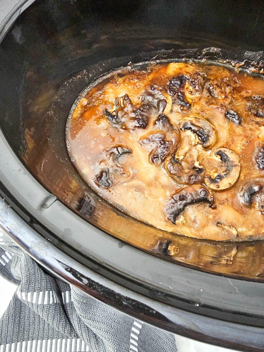 Close-up view of the smothered pork chops and mushrooms submerged in a brown gravy liquid inside the black slow cooker insert after cooking.