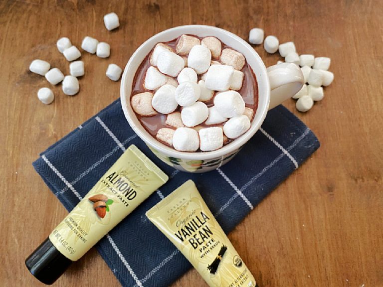 Close up view of homemade hot cocoa flavored with almond and vanilla, served in a white mug and topped with large melting marshmallows. The extract tubes are visible in the foreground.