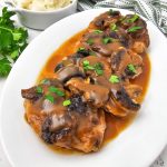 Angled close-up of four tender smothered pork chops topped with thick mushroom brown gravy and parsley on a white platter, with a small dish of mashed potatoes visible.