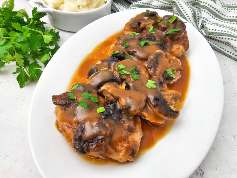 Angled close-up of four tender smothered pork chops topped with thick mushroom brown gravy and parsley on a white platter, with a small dish of mashed potatoes visible.