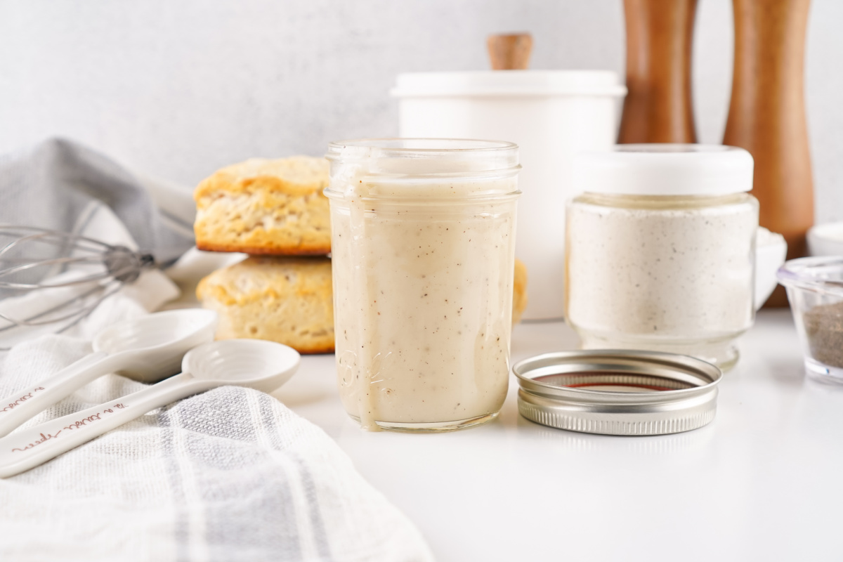 A mason jar of creamy, finished white gravy standing prominently in the foreground, with a jar of the dry mix and a stack of biscuits blurred in the background.