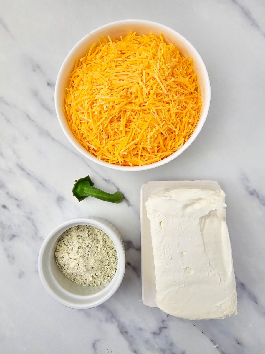 Overhead view of the four main ingredients for the pumpkin cheese ball laid out on a white marble surface: a white bowl of shredded orange cheddar cheese, a block of cream cheese on a small white tray, a small white ramekin with green ranch seasoning mix, and a green bell pepper stem.