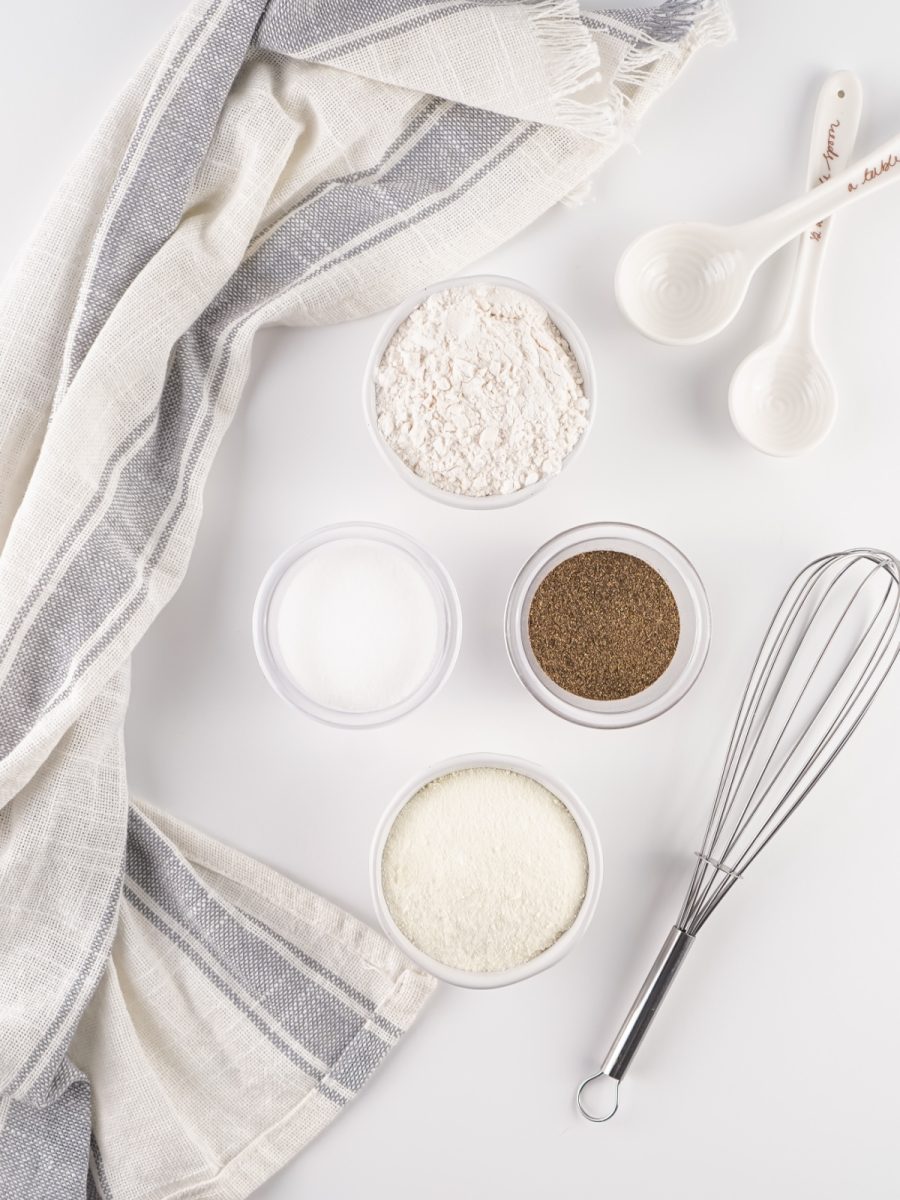 Overhead flat lay of the four dry ingredients: flour, dry milk powder, salt, and black pepper, each in a small bowl, next to a whisk and measuring spoons.