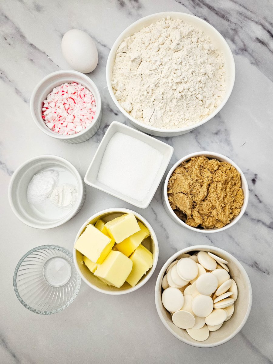 Overhead flat lay of all the ingredients needed, including flour, butter, sugars, egg, crushed peppermint, and white chocolate wafers.