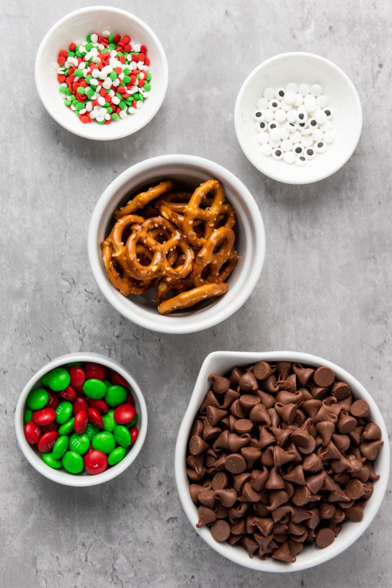 An overhead photo of the required ingredients laid out in white bowls: milk chocolate chips, mini pretzels, red and green M&M's, candy eyes, and festive sprinkles.