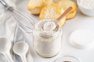 Angled close-up of a wooden spoon scooping out the dry white country gravy mix from a clear jar, with a whisk and biscuits in the background.
