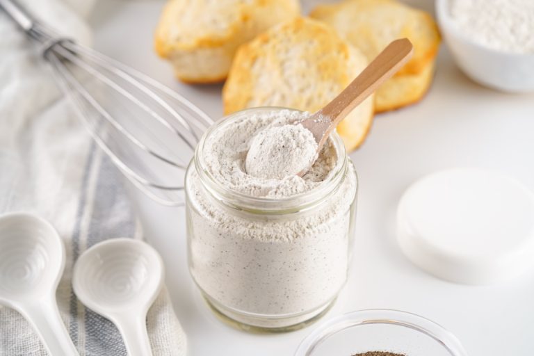 Angled close-up of a wooden spoon scooping out the dry white country gravy mix from a clear jar, with a whisk and biscuits in the background.