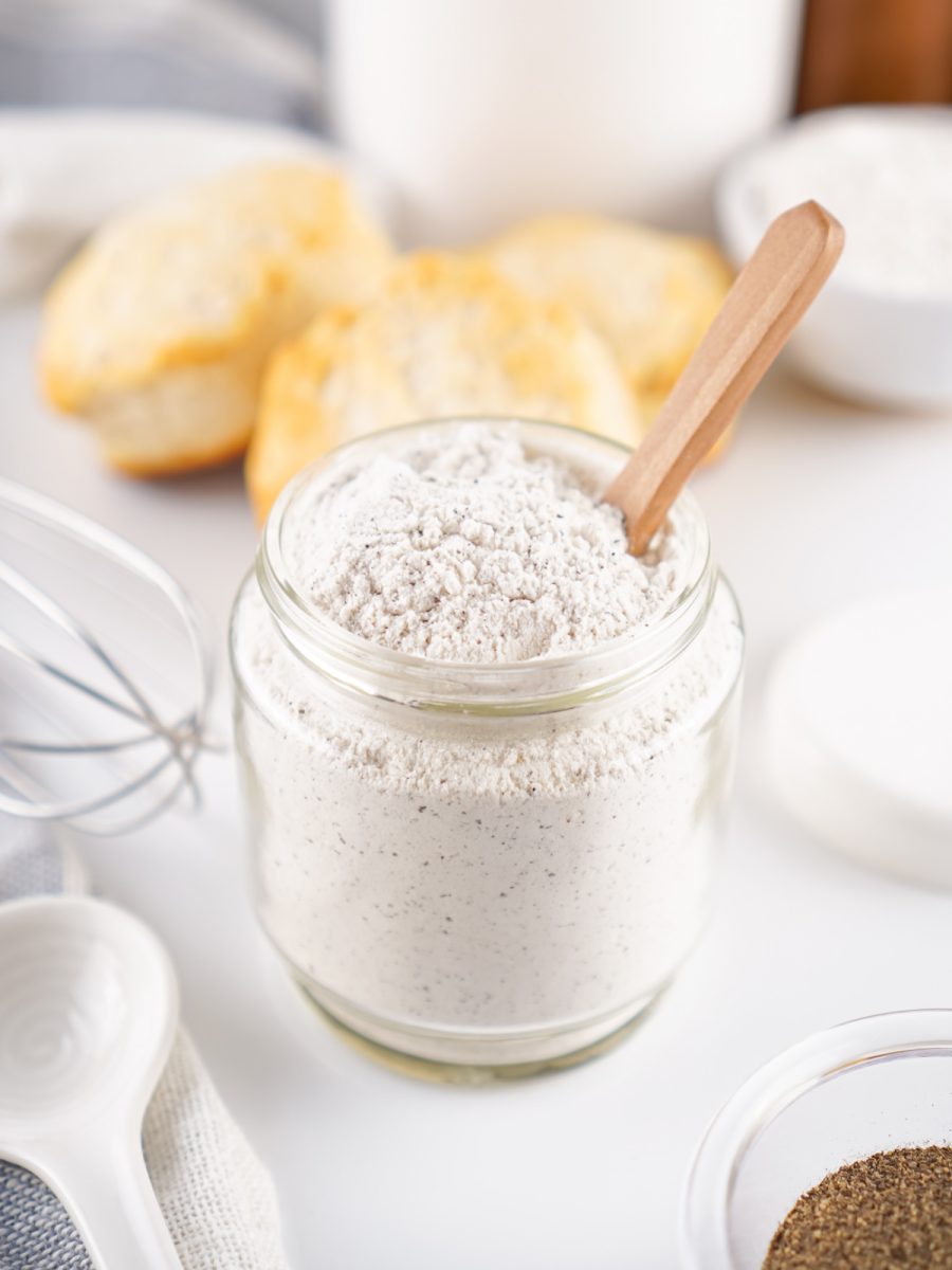 A clear jar filled with the homemade white gravy mix, with a wooden spoon resting inside. Fluffy biscuits and a whisk are visible in the soft background.